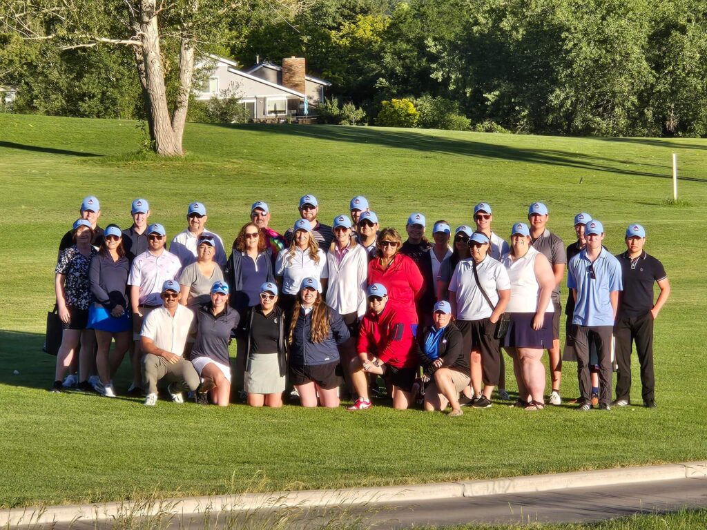 A group of around 30 people wearing hats pose together on a green golf course, with trees and a house in the background. The group is facing the camera under sunny weather.