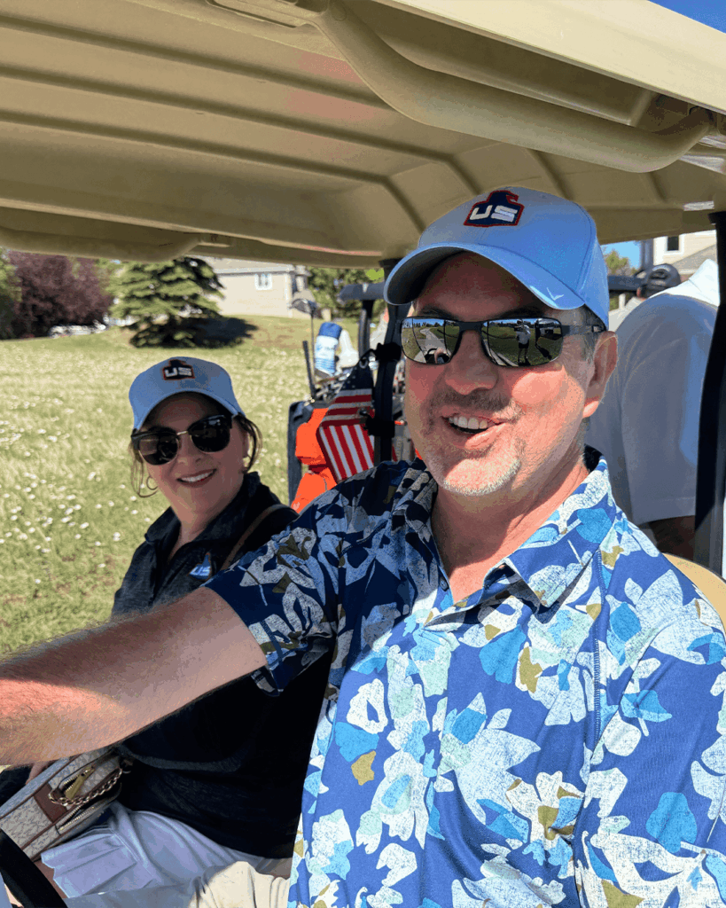 Two adults wearing light blue baseball caps and sunglasses sit in a golf cart on a sunny day. The man in front wears a blue floral shirt; the woman behind him is dressed in black. There is grass and a house in the background.