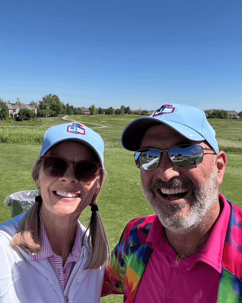 Two people wearing blue caps and sunglasses smile at the camera on a sunny golf course. The man wears a colorful shirt and the woman wears a white jacket with pink underneath. Houses and trees are visible in the background.