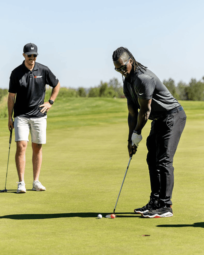 Two men are on a golf course; one is putting while the other stands nearby watching. Both are wearing golf attire, and the weather appears sunny and clear.