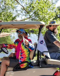 Two men sit in a golf cart near a pond; one wears a colorful tie-dye shirt and laughs while holding a piece of paper. The other wears a gray shirt and hat. A small flag with a red and blue logo is attached to the cart.