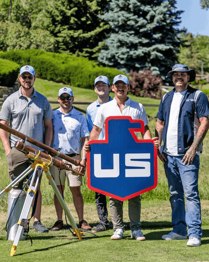 Five men in casual clothing and caps stand outdoors on a golf course, smiling and holding a large shield-shaped sign with US on it. A tripod with equipment is positioned to the left. Trees and grass are visible in the background.