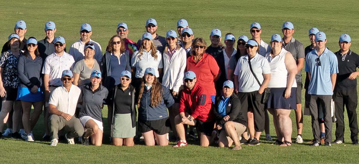 A group of about thirty people pose together on a grassy golf course, all wearing blue baseball caps and casual golf attire, with sunlight casting shadows on the ground.