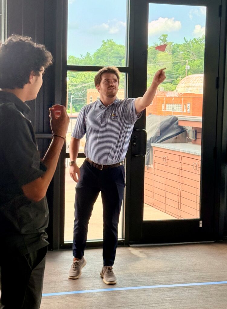 Two men stand indoors near an open door. One man in a striped polo shirt and dark pants is throwing a paper airplane while the other man, in a dark shirt, watches and holds a drink. Outdoor scenery is visible through the door.