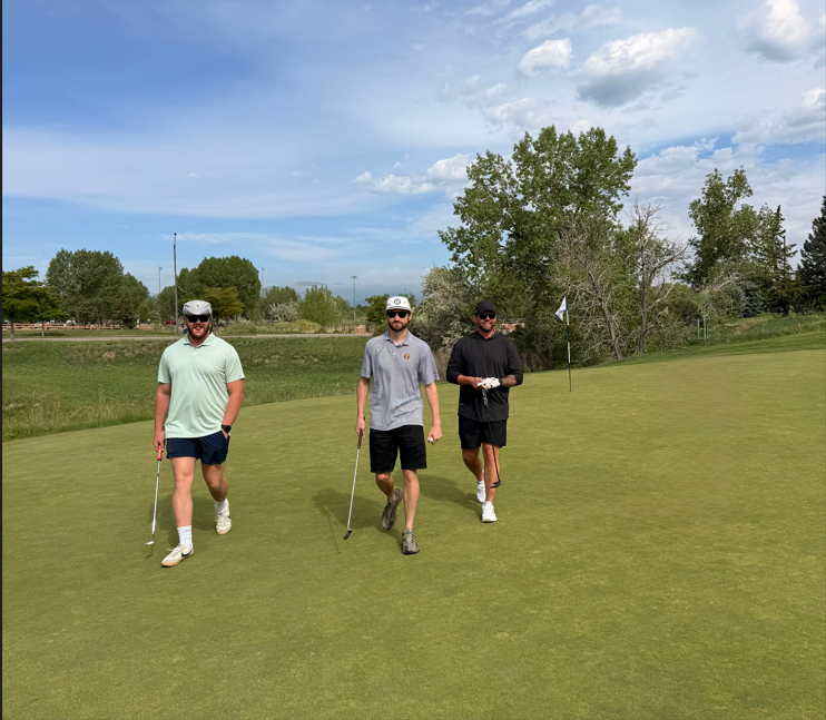 Three people wearing casual golf attire and sunglasses walk on a golf green holding clubs, with a golf flag and trees in the background under a partly cloudy sky.