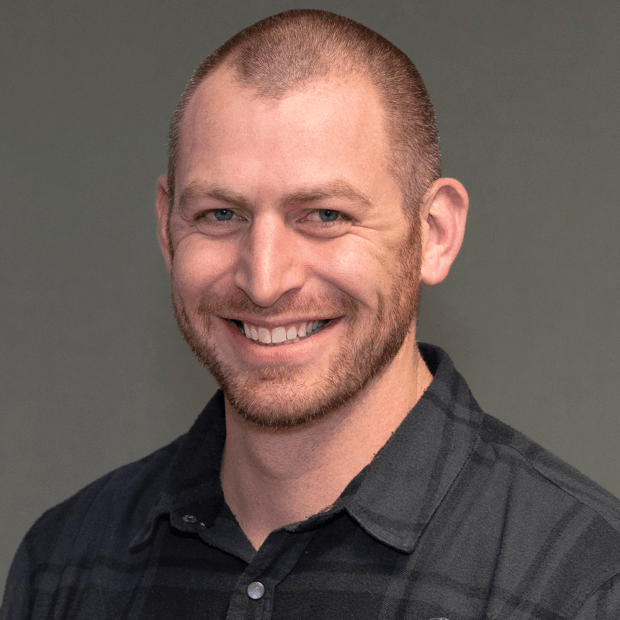 A man with a shaved head and beard is smiling at the camera. He is wearing a dark plaid shirt and is posed against a plain gray background.