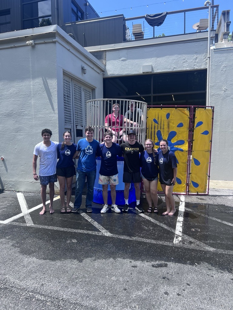 Eight people stand in front of a dunk tank outside on a sunny day. One person is sitting inside the dunk tank, slightly elevated above the group. The ground and part of the group are wet.