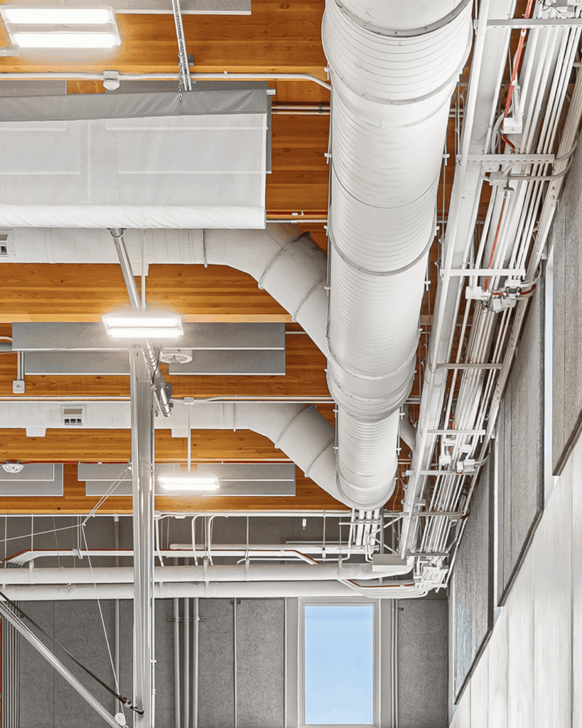 Exposed pipes, ducts, and wiring run along a high ceiling with wood panels and acoustic coverings. Bright lighting fixtures hang from the ceiling; a window and part of a wall are visible on the right.