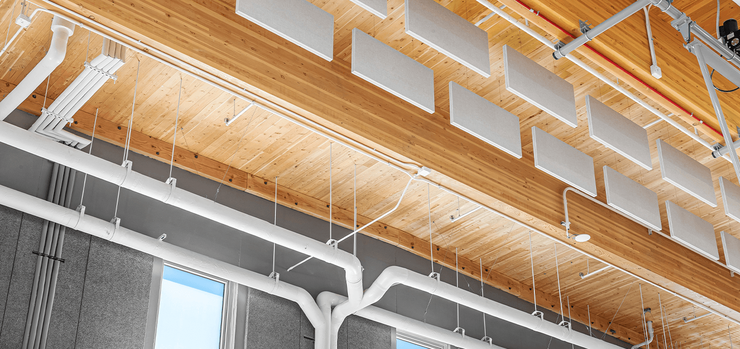 Wooden ceiling with suspended rectangular acoustic panels, white pipes, and exposed ducts in a modern building. Gray walls and large windows are partially visible at the bottom of the image.