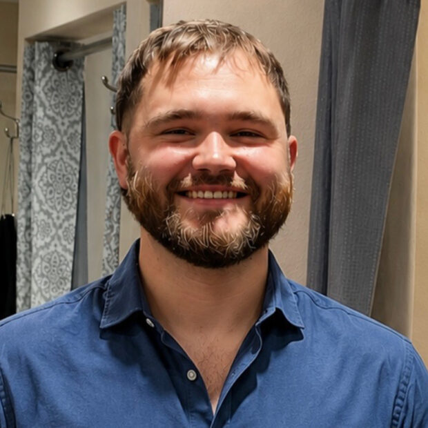 A man with short hair and a beard is smiling, wearing a blue button-up shirt. He is standing indoors with patterned and solid curtains visible in the background.