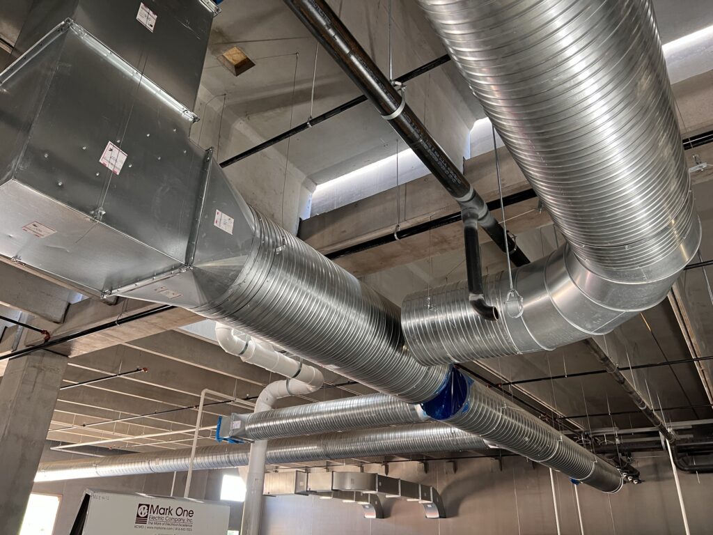 Large silver HVAC ducts and rectangular metal air vents run across the ceiling of an unfinished building, with exposed pipes and concrete columns visible in the background.