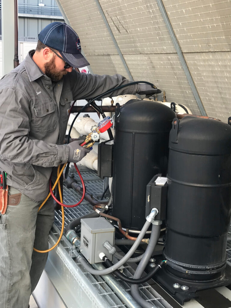 A man wearing a cap, sunglasses, work gloves, and a gray uniform uses a gauge and hose to service or inspect industrial HVAC equipment on a rooftop.