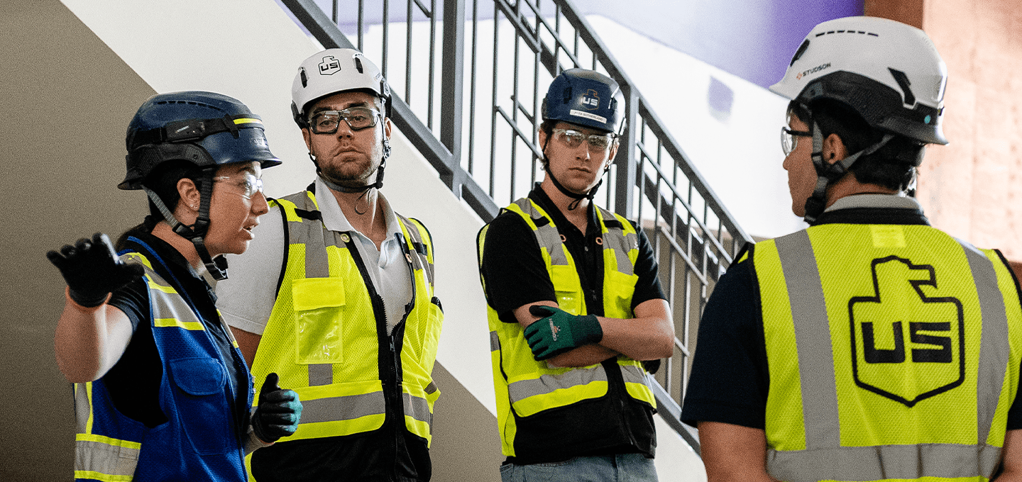 Four people wearing safety vests, helmets, and protective eyewear stand indoors, near a stairwell, engaged in conversation. One person gestures with their hand while the others listen.