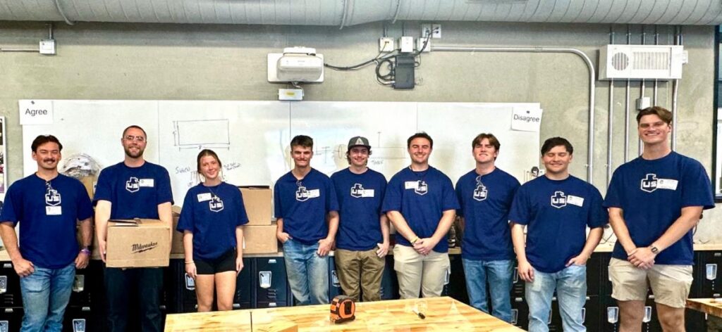 Nine people wearing matching blue shirts stand in a workshop or classroom with whiteboards behind them. One person holds a cardboard box. Tables and tools are visible in the foreground.