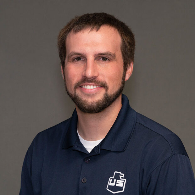 A man with short brown hair, a beard, and a mustache is smiling at the camera. He is wearing a navy blue collared shirt with a white logo on the left side, in front of a plain gray background.