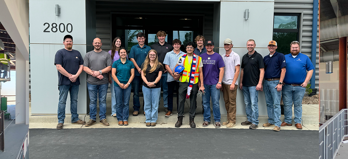 Fifteen people stand in a row outside a building with the number 2800 visible on the wall. One person wears a safety vest and hard hat. The group poses on a paved area in front of glass doors.