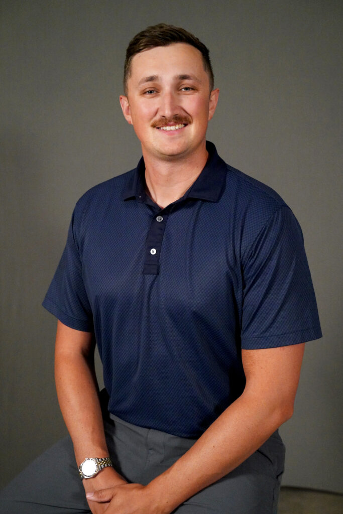 A man with short brown hair and a mustache sits against a plain gray background. He is wearing a dark blue short-sleeved polo shirt, gray pants, and a watch, and is smiling at the camera.