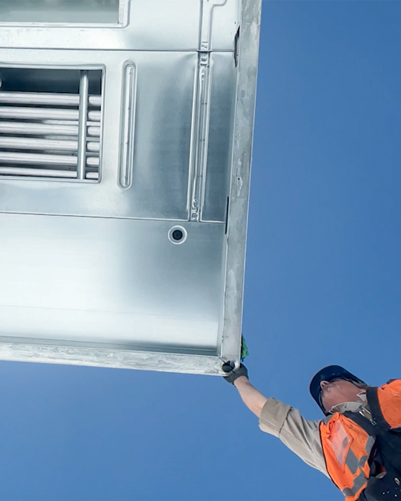 A worker in an orange vest and hard hat steadies a large metal unit suspended above, with a clear blue sky in the background.