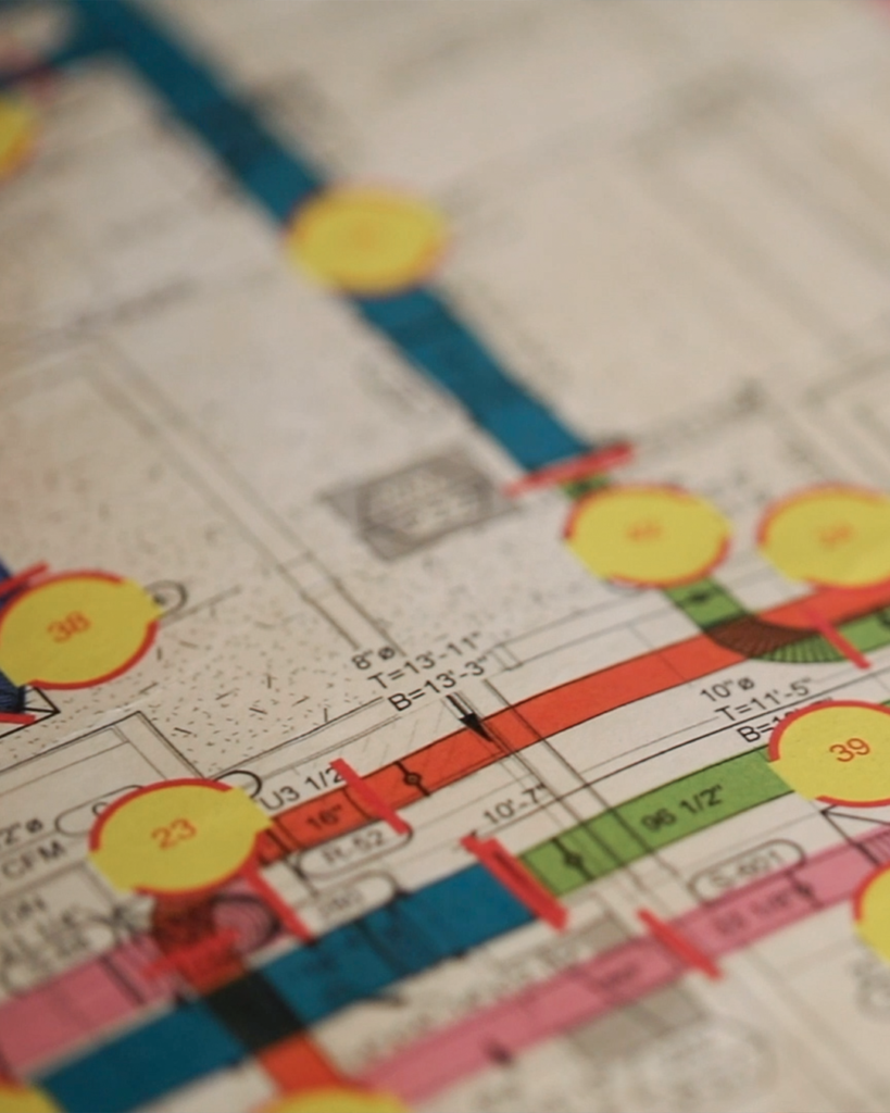 A close-up of a technical drawing or blueprint with colored lines and yellow numbered circles, possibly representing a buildings wiring or piping layout. The image is slightly out of focus.