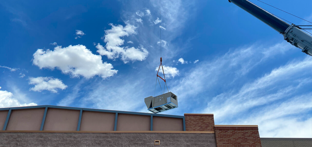 A crane lifts a large HVAC unit above the roof of a commercial building under a blue sky with scattered clouds.
