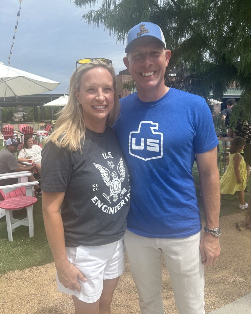 Two adults stand outdoors and smile at the camera. The woman wears a dark U.S. Engineers t-shirt and white shorts. The man wears a blue t-shirt with a white logo and a blue cap. People and picnic tables are in the background.