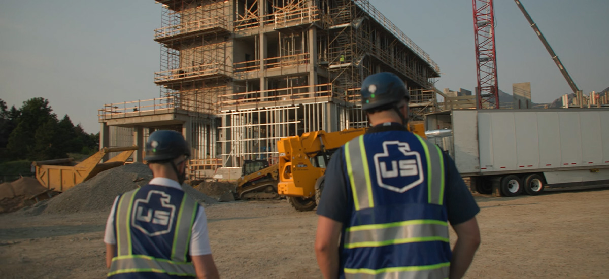 Two construction workers in safety vests and helmets face a multi-story building under construction with scaffolding, a yellow machine, and a semi-truck nearby.