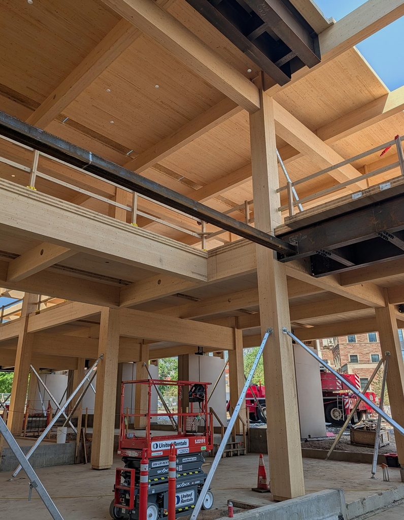 Partially constructed building with exposed wooden beams, scaffolding, and construction equipment including a scissor lift and traffic cones. Structural supports and unfinished flooring are visible throughout the site.