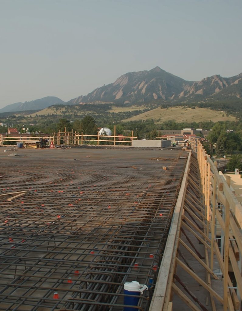 A construction site with a large, flat surface covered in rebar, wooden supports along the edge, and mountains with green hills in the background under a clear sky.