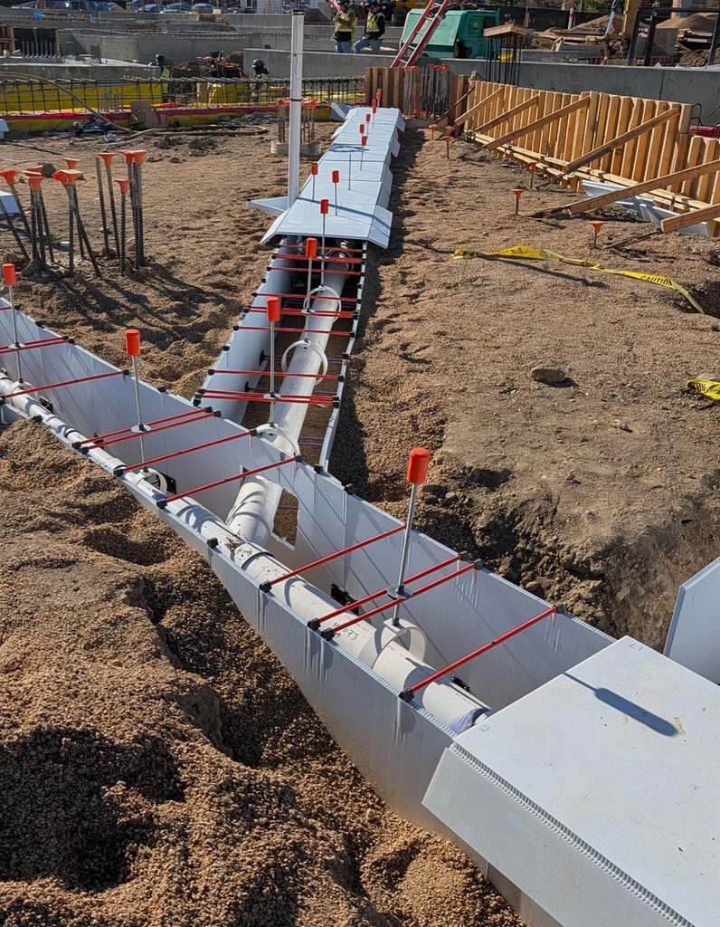 A construction site shows plumbing pipes laid in trenches surrounded by gravel and temporary formwork. Red safety caps are on vertical rods, and wooden framing with caution tape is visible in the background.