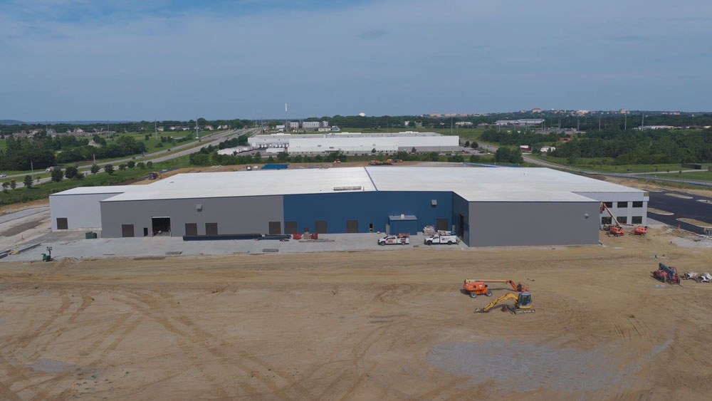 A large, rectangular industrial warehouse with a white roof, gray and blue exterior walls, and construction equipment in front. The site is surrounded by open land and other buildings, with trees in the background.