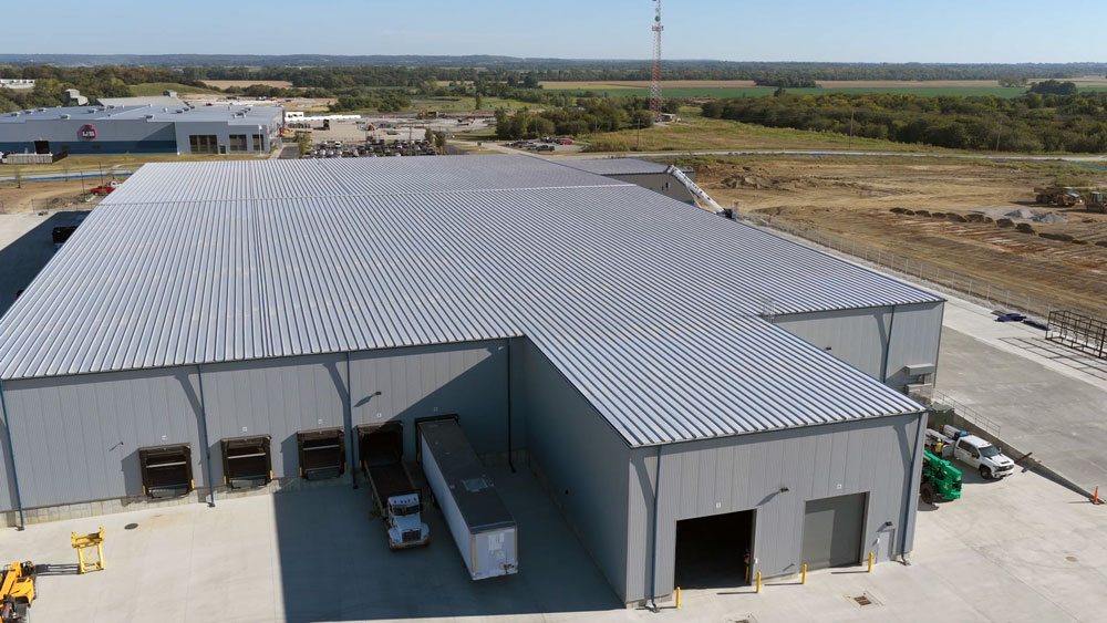 Large industrial warehouse with a grey metal roof and multiple loading docks. A white truck trailer is backed up to one dock. The building is surrounded by concrete and situated in a mostly rural area with open fields.