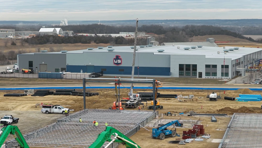 Construction site with steel framework and machinery in the foreground. Several workers are visible. Behind the site is a large industrial building with the letters US on the wall, surrounded by a rural landscape and overcast sky.