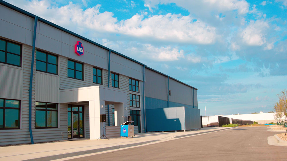 A modern industrial building with a gray facade, large windows, and the US logo above the entrance, under a blue sky with some clouds. The area outside is paved and mostly empty.