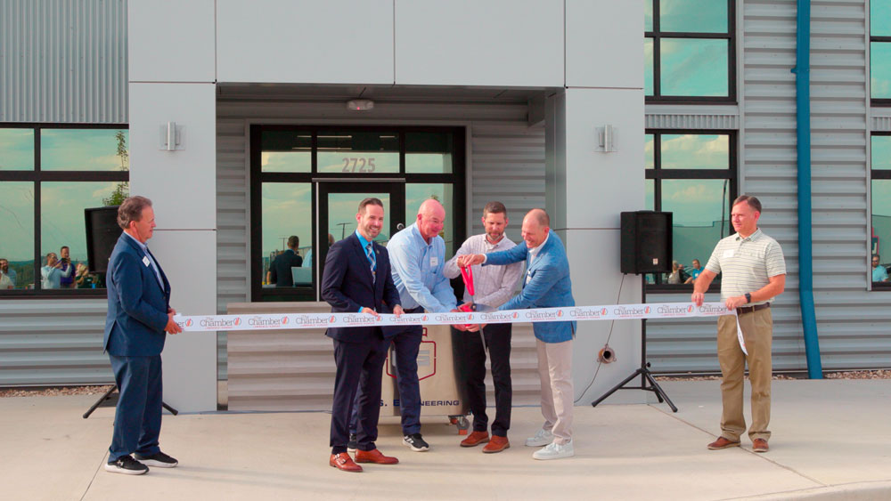Six men stand in front of a building for a ribbon-cutting ceremony. Four men in the center hold scissors to cut the ribbon, while two men stand on either side watching. The building entrance is visible in the background.