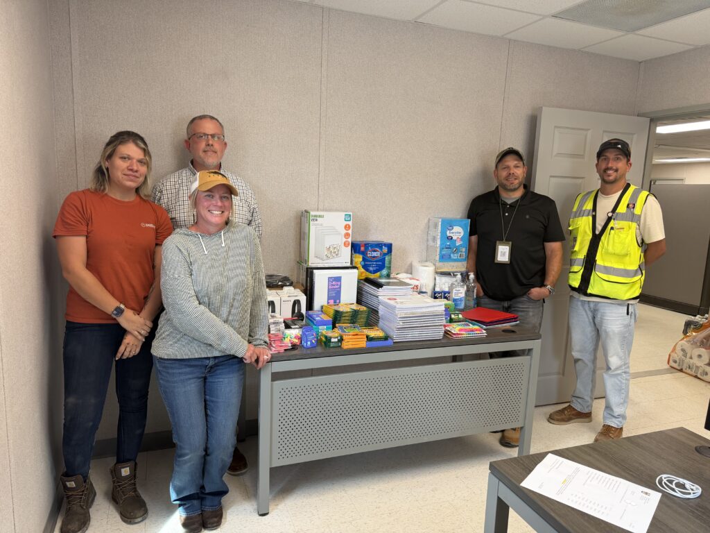 Five adults stand behind a table filled with assorted school supplies, notebooks, and snacks in an office setting. The group appears to be posing for a photo, and the room has light-colored walls and a closed door.