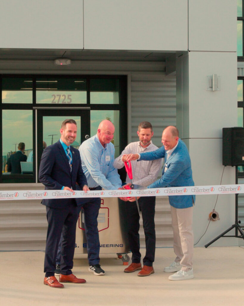 Four men in business attire stand in front of a building while two of them cut a ceremonial ribbon; the ribbon displays the word Chamber and the building address 2725 is visible above the door.